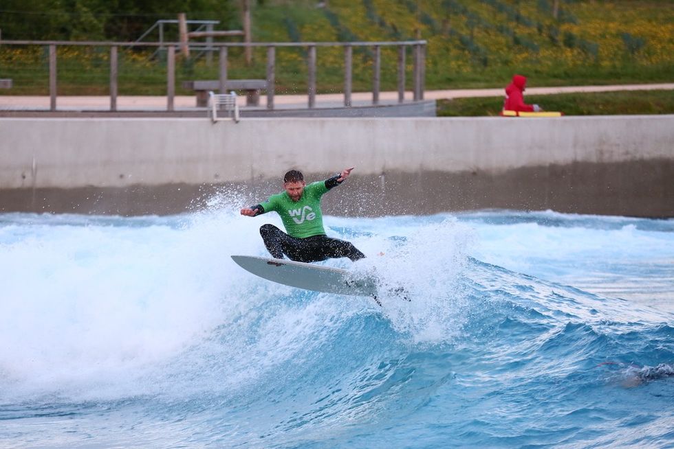 Surfer in green shirt riding a wave at a wave pool, with a person in red sitting nearby.