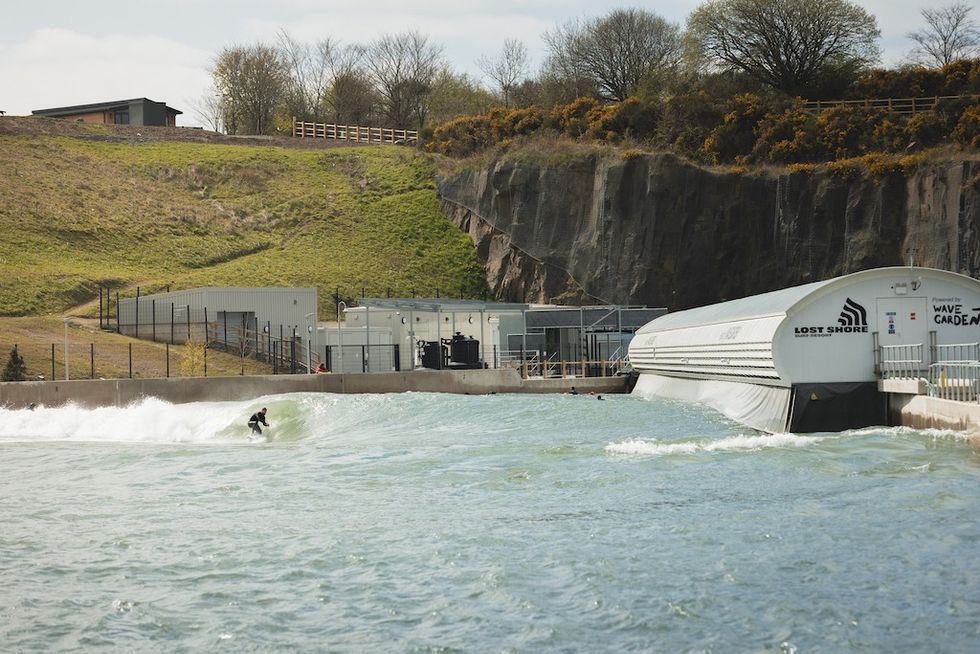 Surfer rides wave in an artificial surfing pool near a rocky hillside.