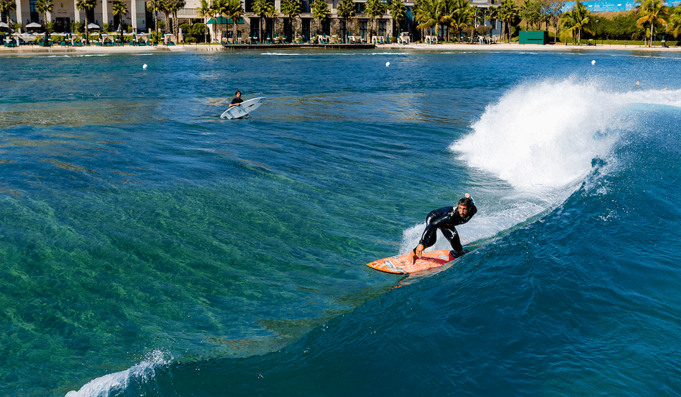 Surfer riding a clear blue ocean wave near the shoreline with palm trees in the background.