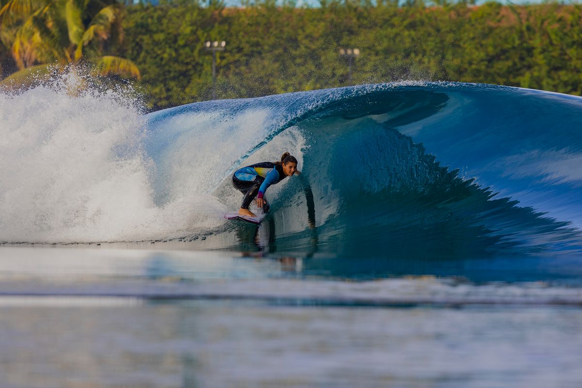 Surfer riding a perfect, curling blue wave with lush greenery in the background.