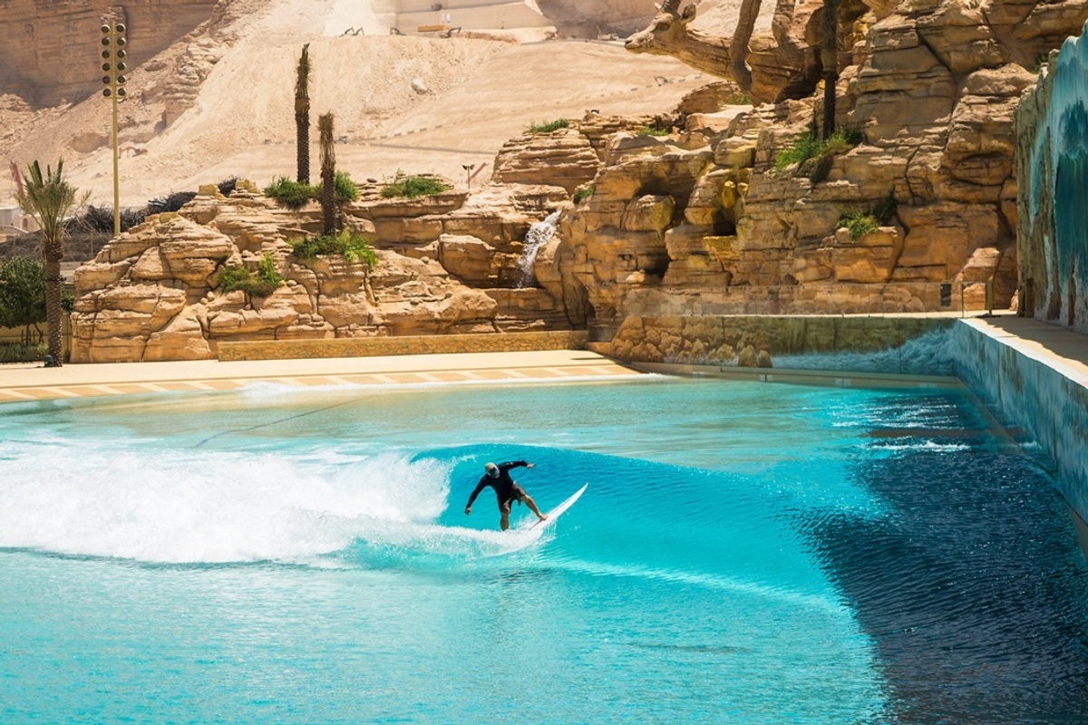 Surfer riding a wave in a blue pool, with rocky desert backdrop and palm trees.