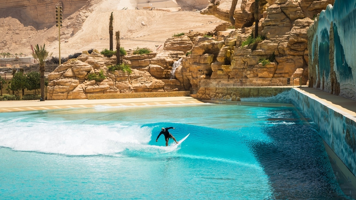 Surfer riding a wave in a blue pool, with rocky desert backdrop and palm trees.
