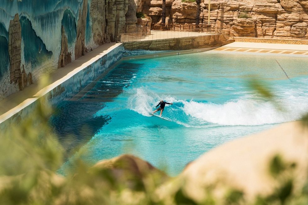 Surfer riding a wave in a large, blue wave pool with rocky landscape and mural backdrop.
