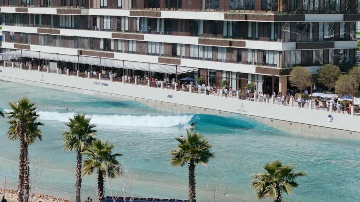 Surfer riding a wave in a pool beside a modern building and palm trees.