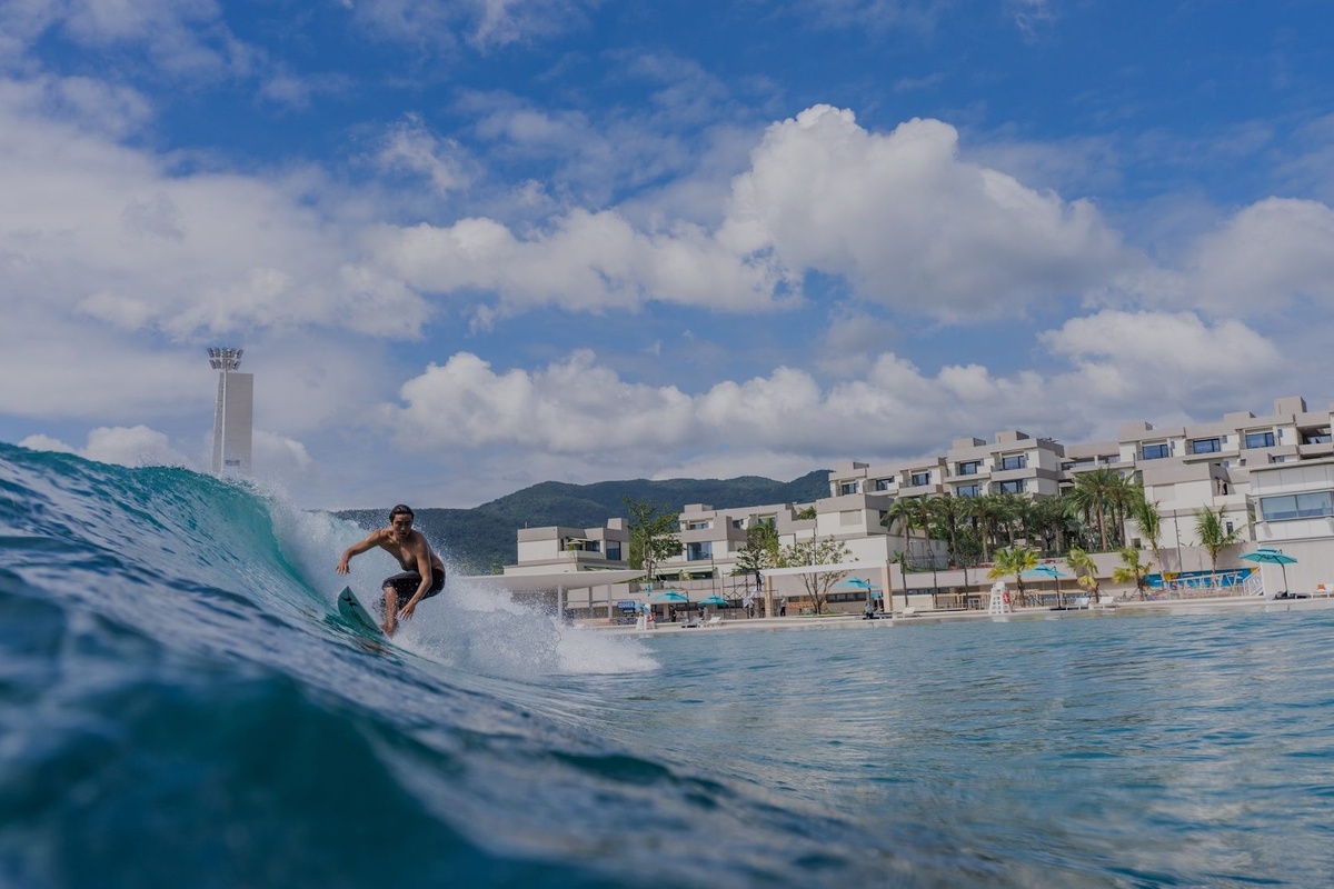 Surfer riding a wave near modern beachfront buildings under a partly cloudy blue sky - Riyue Bay Surf Resort