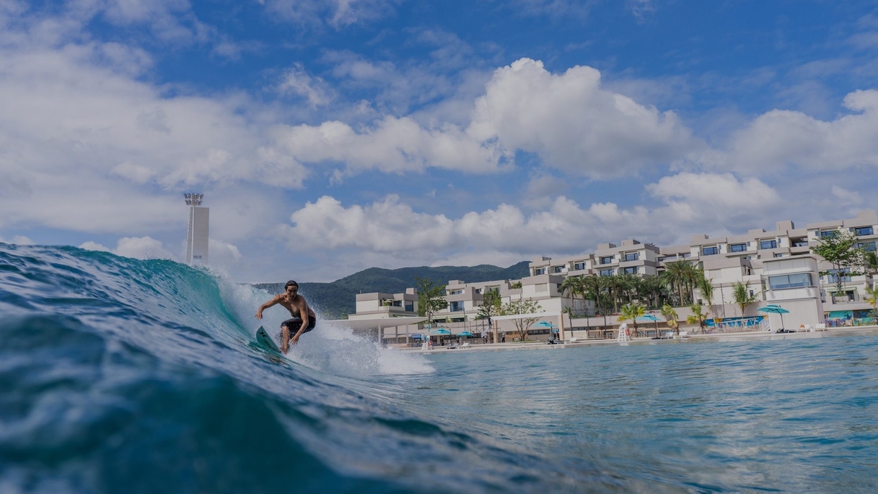 Surfer riding a wave near modern beachfront buildings under a partly cloudy blue sky - Riyue Bay Surf Resort