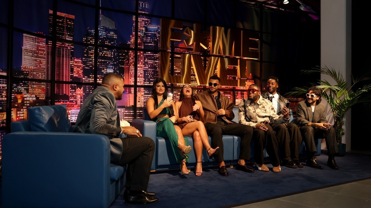 Talk show host with six guests laughing on a blue couch, cityscape backdrop.