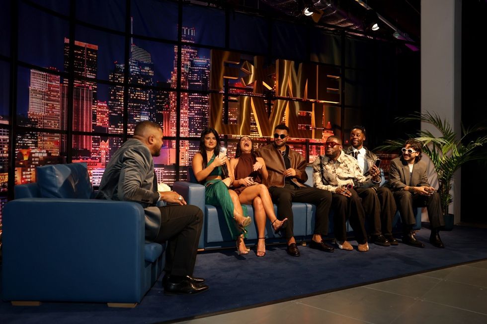 Talk show host with six guests laughing on a blue couch, cityscape backdrop.