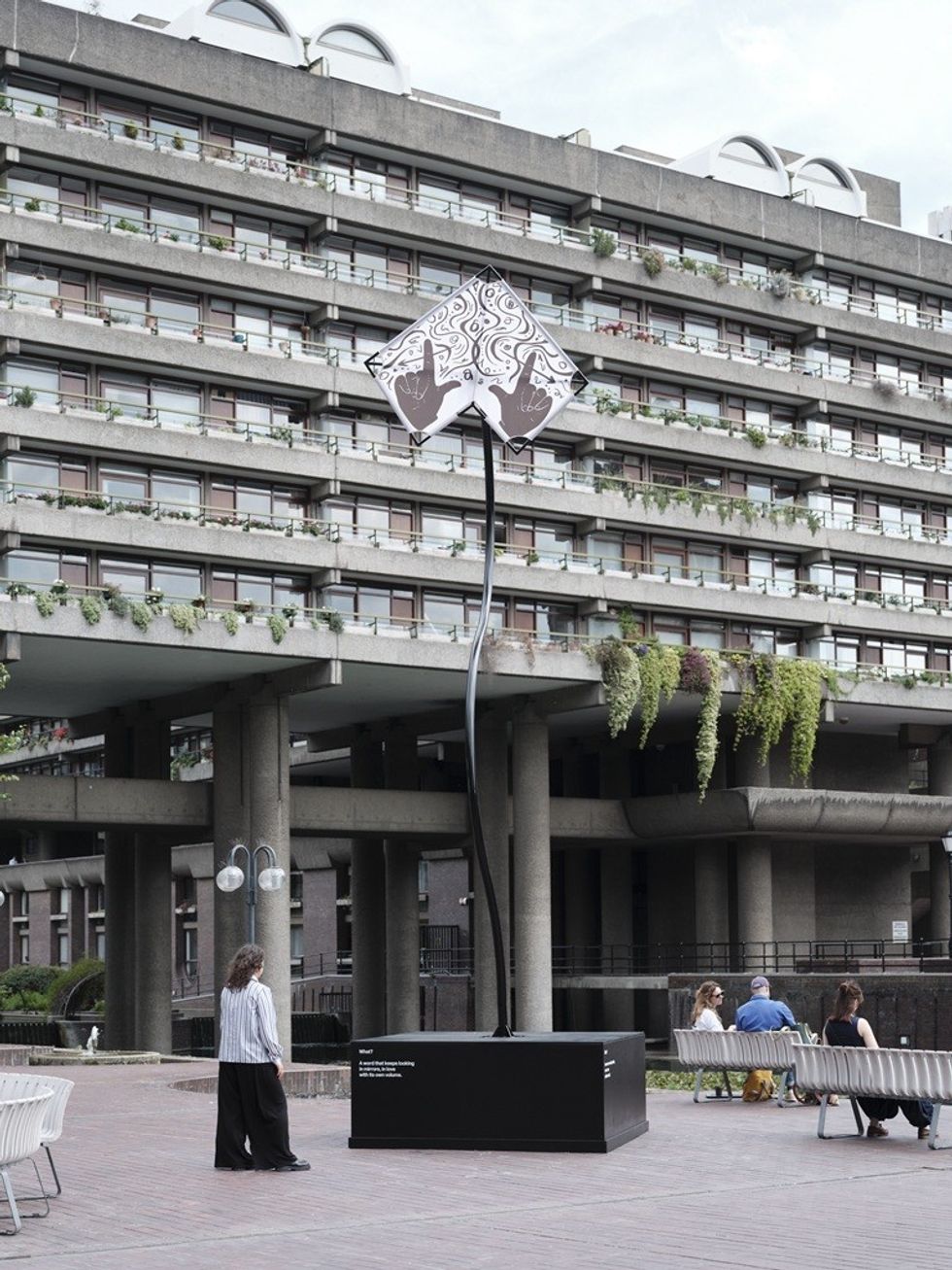 Tall sculpture with intricate design in front of a modern, concrete building.