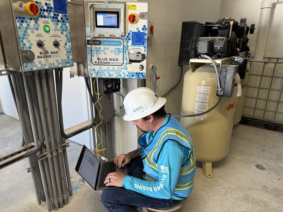 Technician in hard hat using a laptop beside an electrical panel in an industrial setting.