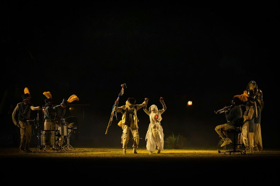 The Cort\u00e8ge: A Festive Funeral for Our Times Performers in colorful costumes on stage, surrounded by musicians in dim lighting.