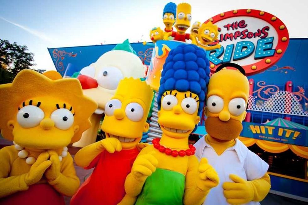 The Simpsons family in costumes at an amusement park ride entrance.