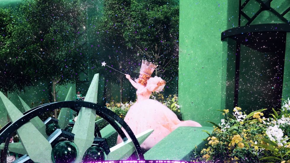 The Wizard of Oz scene with a magical figure in a pink gown and wand amid greenery and sparkles.