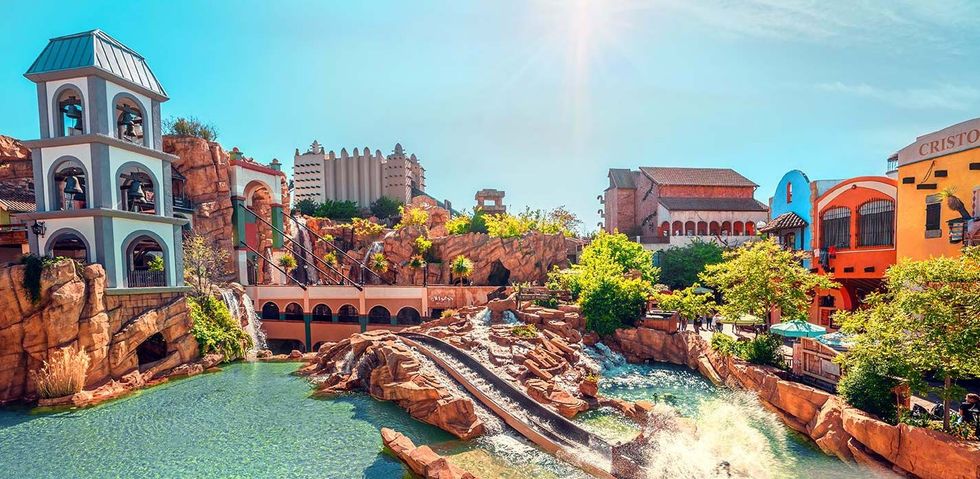 Theme park scene with river rapids, colorful buildings, and bright sunlight.