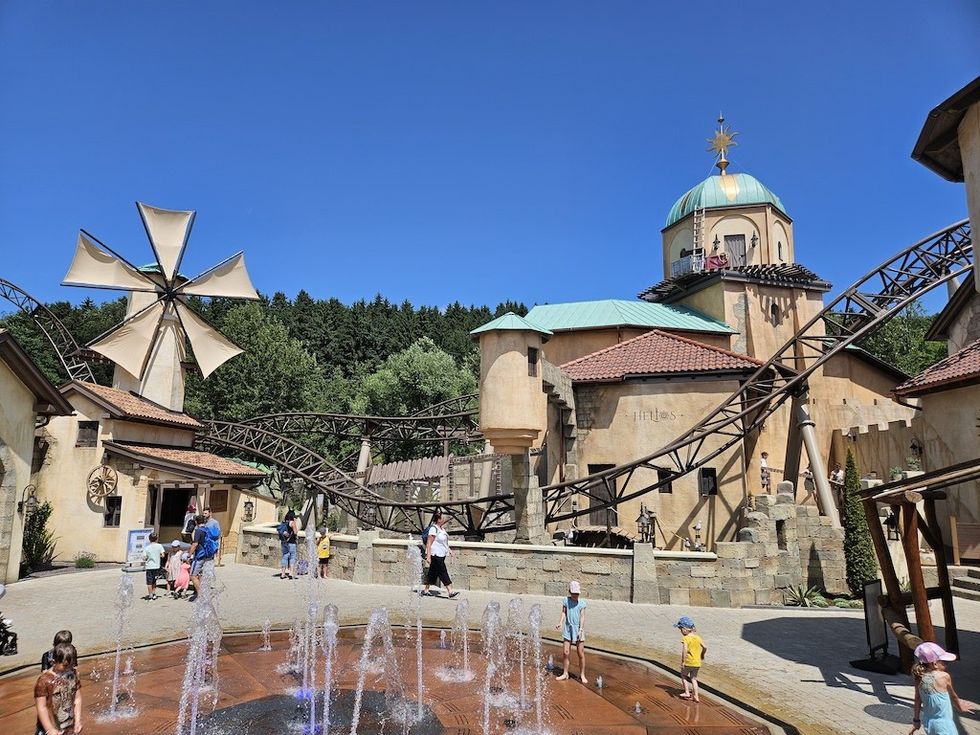 Theme park with a roller coaster, windmill, and fountains, under a clear blue sky.