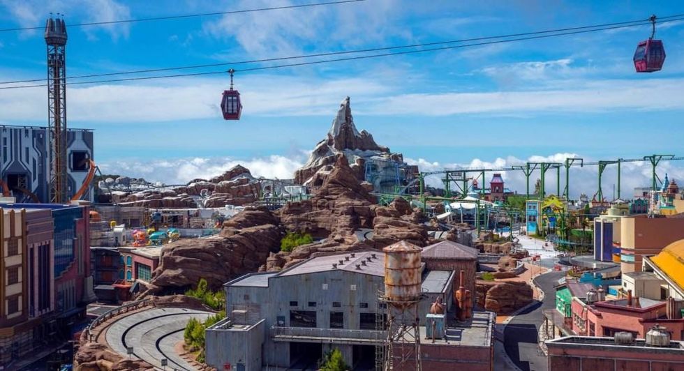 Theme park with rocky mountain, buildings, and cable cars under a blue sky.