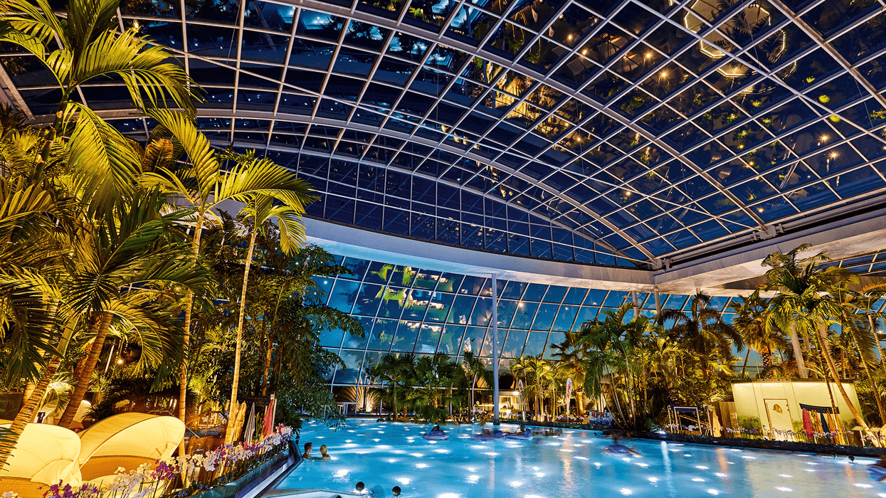 Therme Euskirchen's indoor tropical pool under a glass dome, surrounded by palm trees at night.