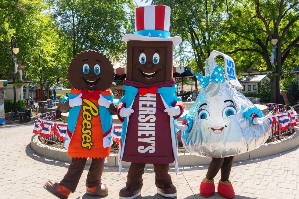 Three chocolate mascots posing in a sunny, decorated setting at Hersheypark