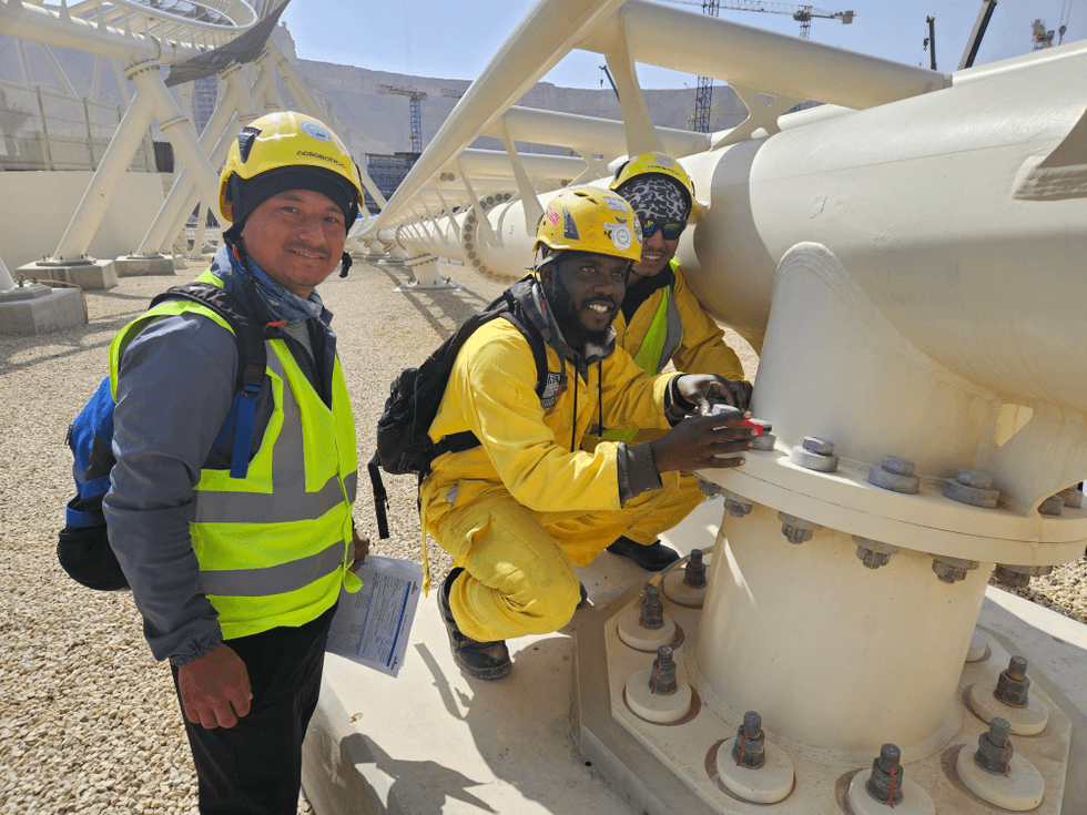 Three construction workers inspect large industrial piping outdoors.