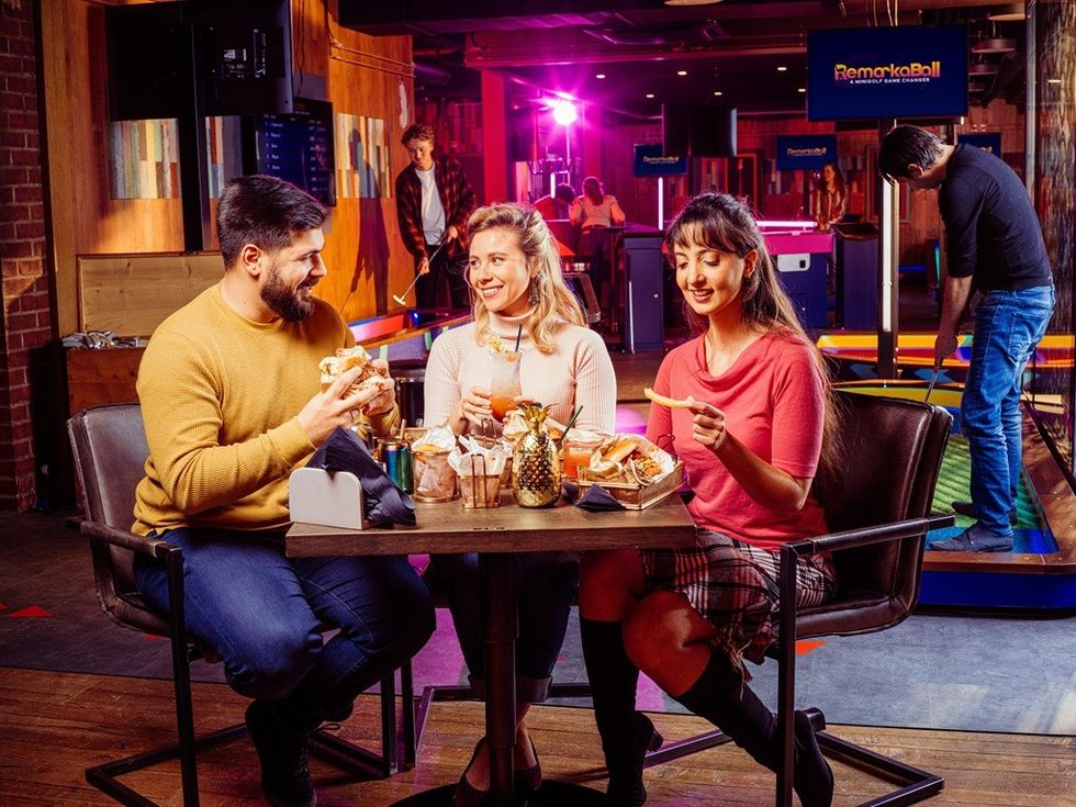 Three friends dining and chatting in a vibrant, neon-lit indoor minigolf venue.