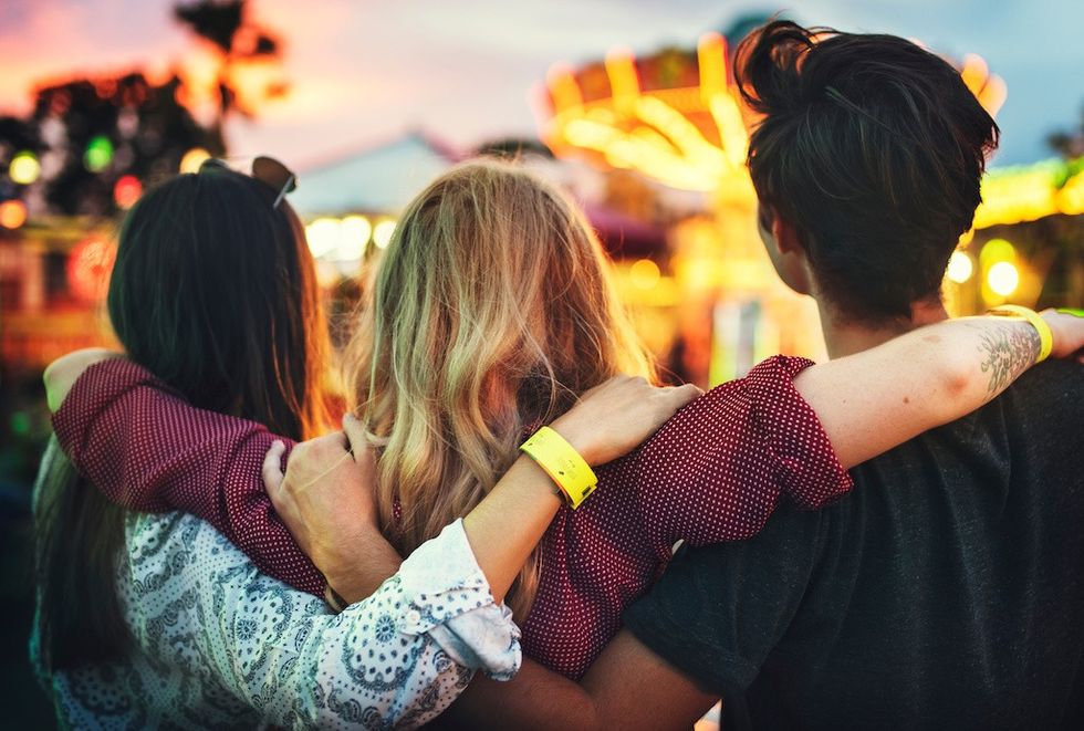 Three friends with arms around each other, looking at a lit-up amusement park ride at dusk.