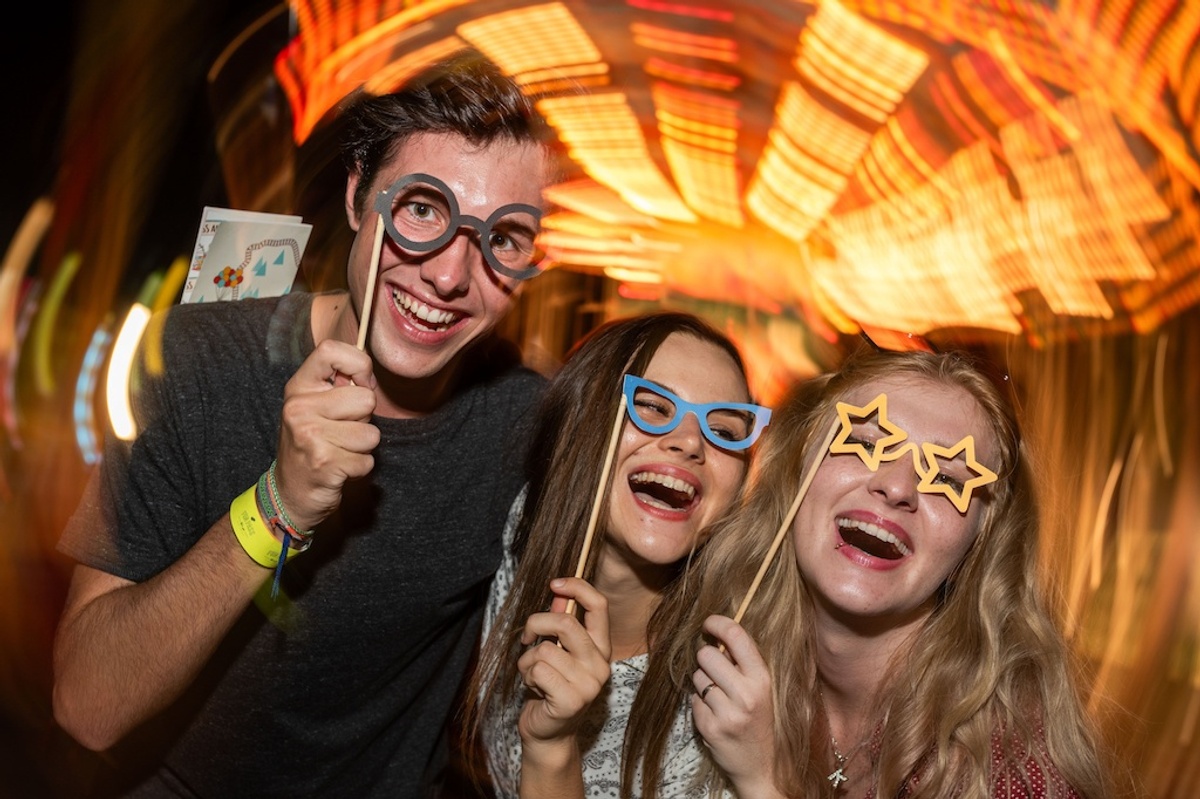 Three friends with novelty glasses laugh amidst bright, swirling lights at a funfair.