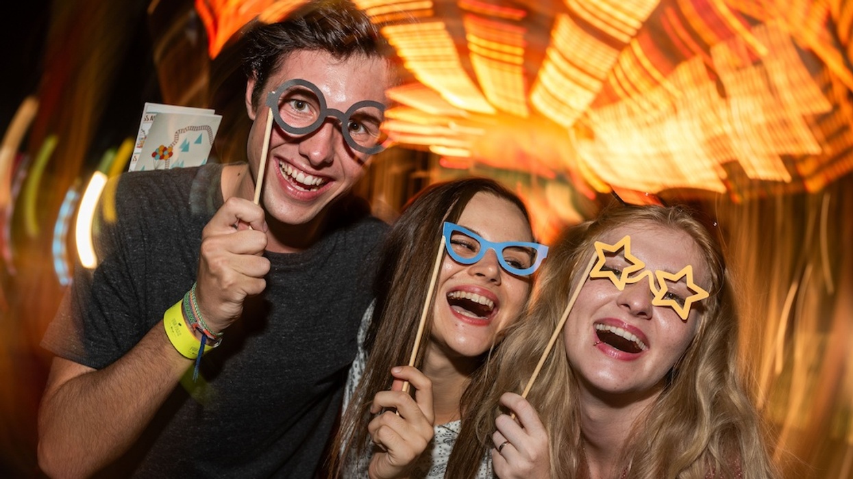 Three friends with novelty glasses laugh amidst bright, swirling lights at a funfair.