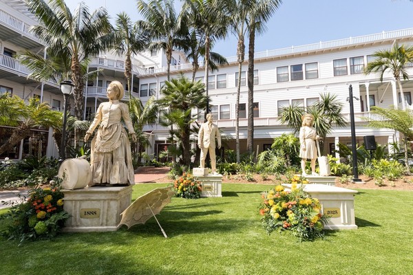 Three human statues in a garden area with palm trees and a white building.