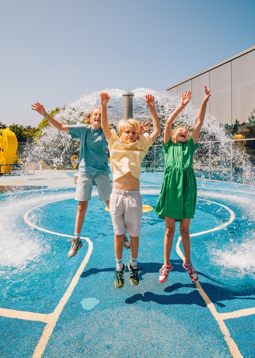 Three kids joyfully jump at a splash pad, surrounded by water spray and sunshine.