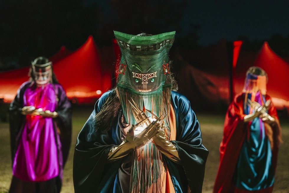 Three masked figures in colorful robes stand before red tents at night.