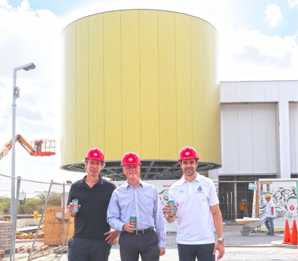Three men (Guus Slabbekoorn, group CFO of MetaCorp (middle), Shensly Tromp, group COO of MetaCorp (right), and Markus Konings, CEO of Sandenburg Experiences) in red hard hats holding cans in front of a large, yellow, cylindrical structure designed to resemble a giant beer tank.