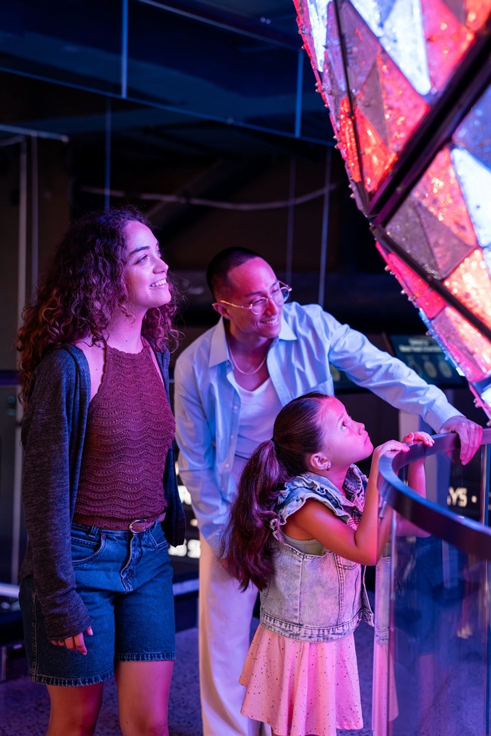Three people admire a large, glowing geometric structure in vibrant lighting in the Times Travel experience.