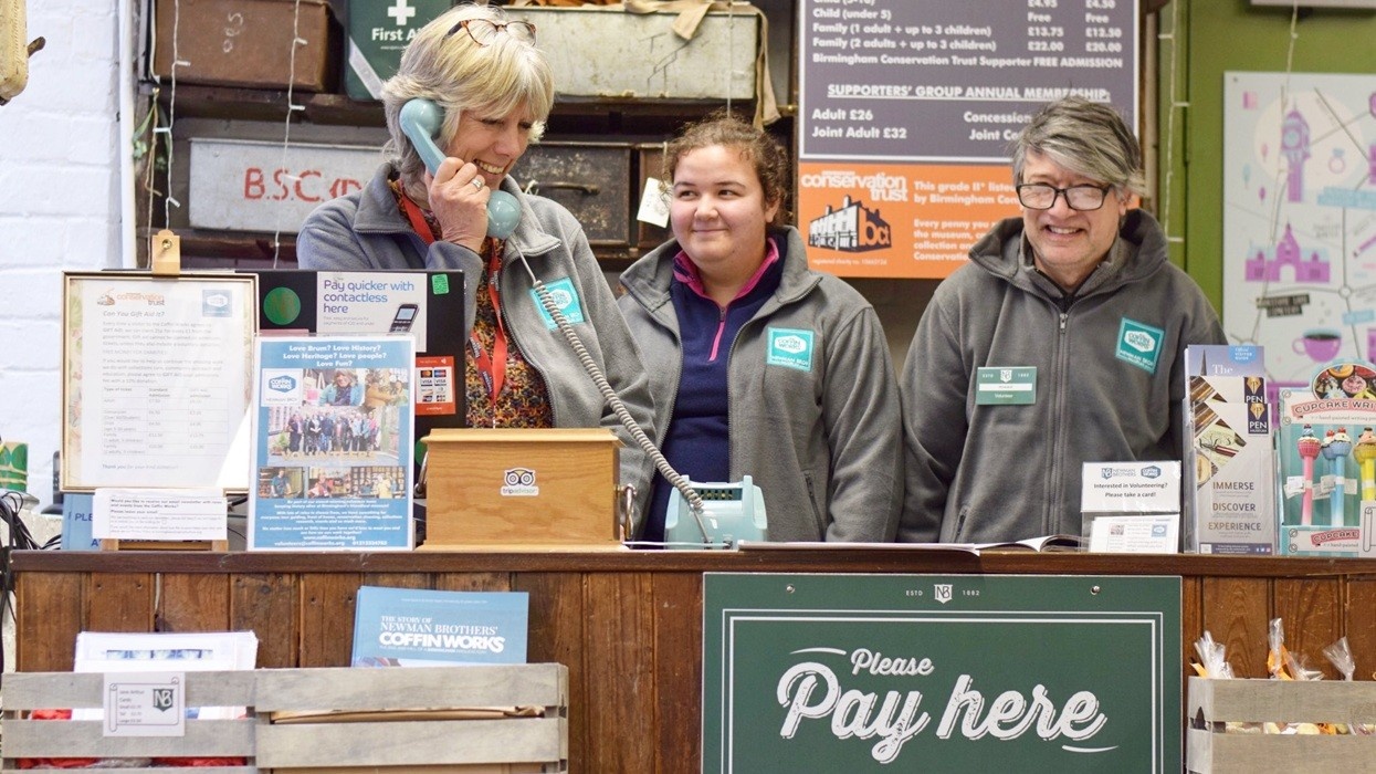 Three people at a wooden counter with a "Please Pay Here" sign, one on the phone.