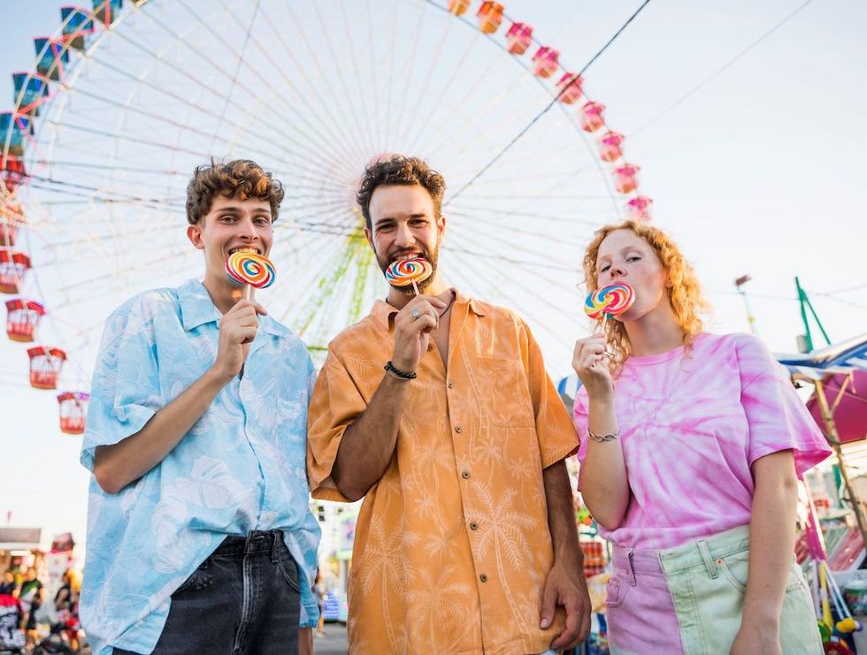 Three people enjoying lollipops in front of a Ferris wheel at a fair.