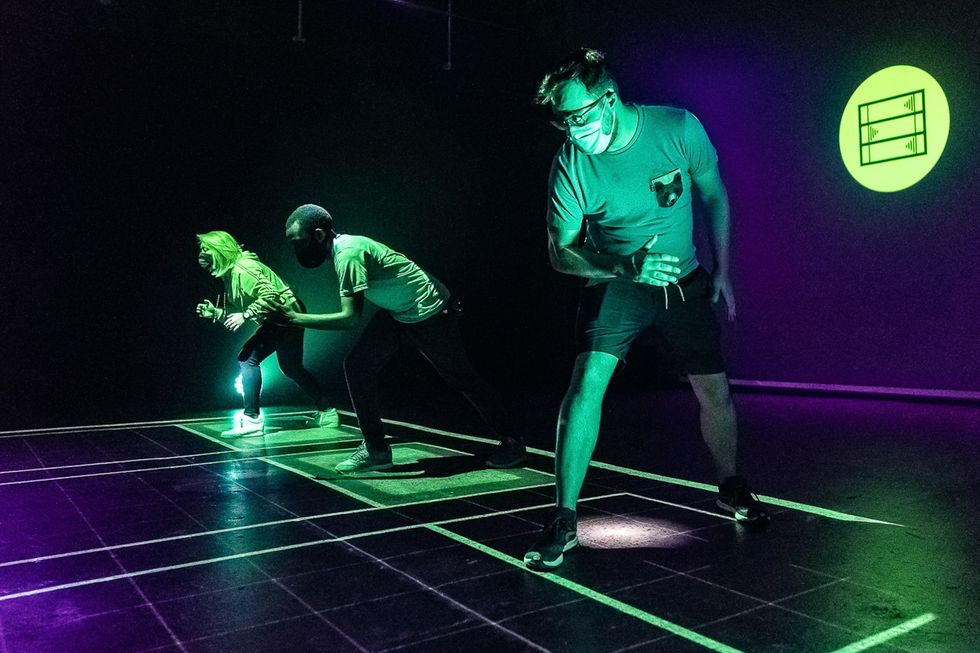 Three people in active poses on a grid, neon lights and projections in the background.