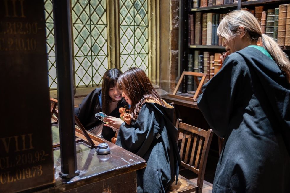 Three people in dark robes, gathered in a library, looking at a phone.