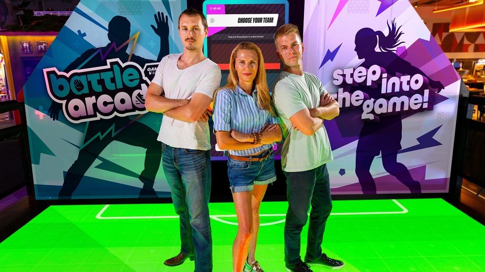 Three people in front of a Battle Arcade game display with colourful graphics.