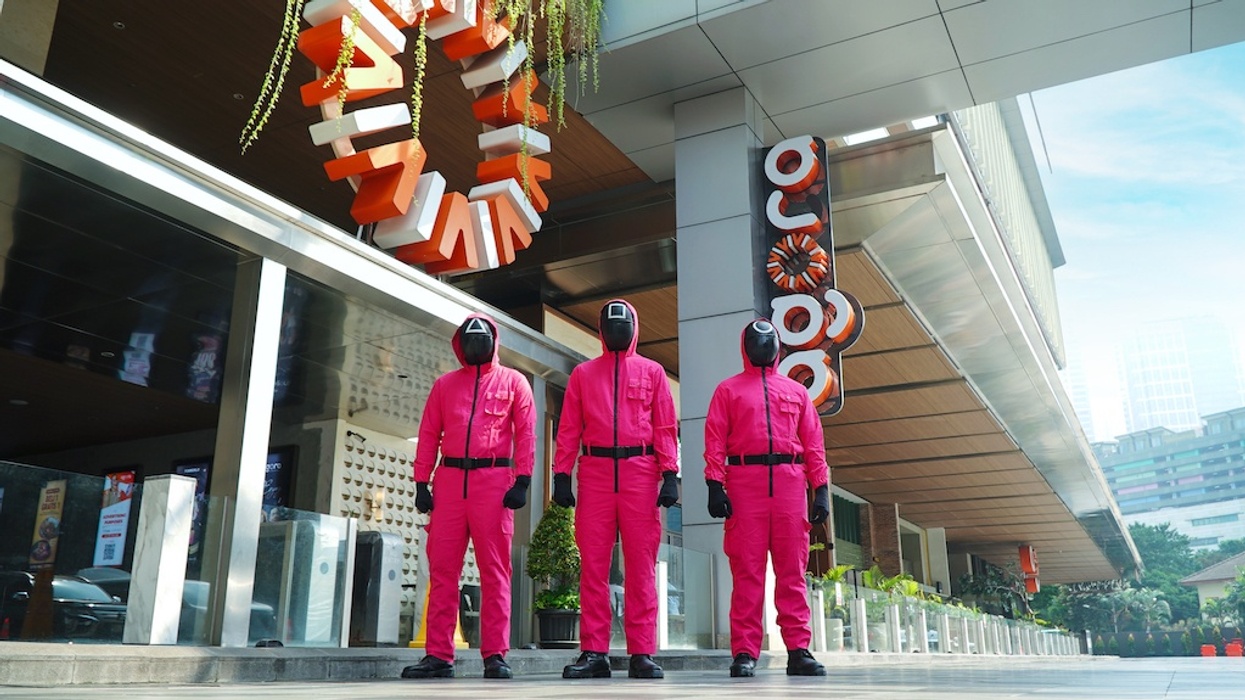 Three people in pink suits and masks from Netflix Squid Game stand outside a modern building, Agora Mall
