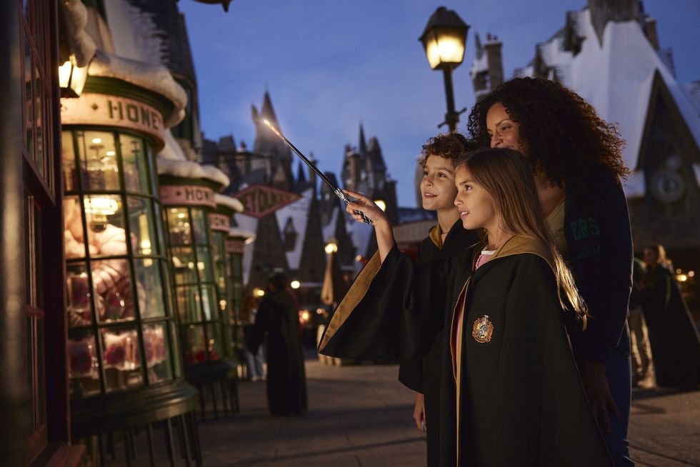 Three people in robes, one with a wand, at Universal's Hogsmead Village at dusk.