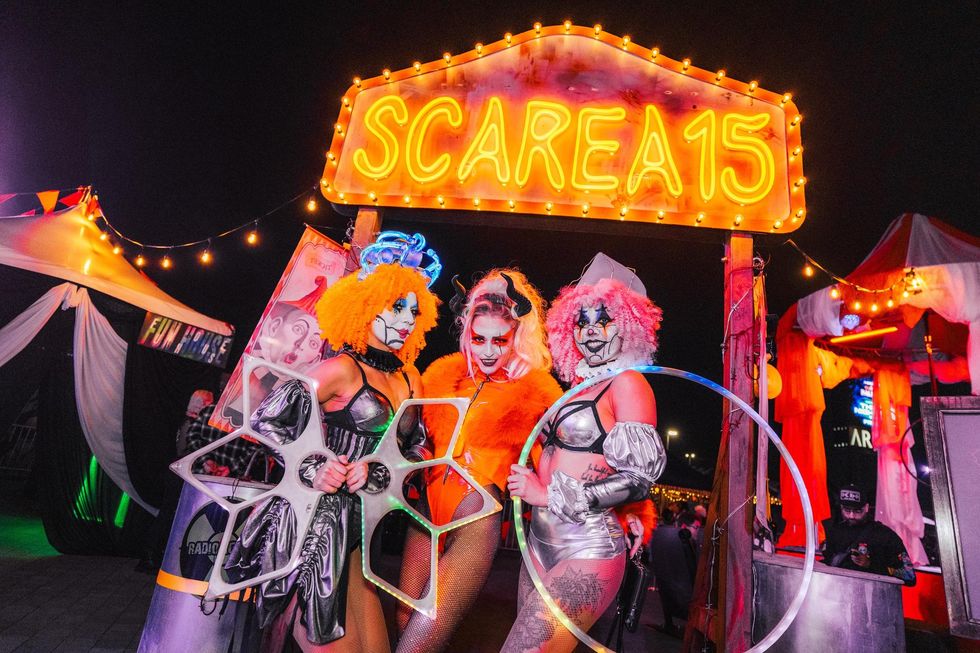 Three people in vibrant costumes under a neon "ScareA15" sign at night.