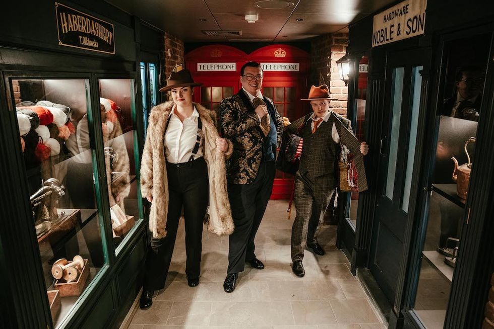 Three people in vintage attire pose in a hall with shop facades and a red phone booth.
