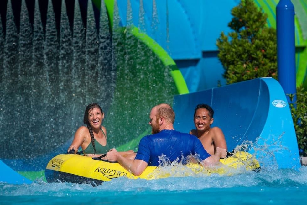 Three people laughing on a water park slide raft under a cascading water curtain at Aquatica Orlando