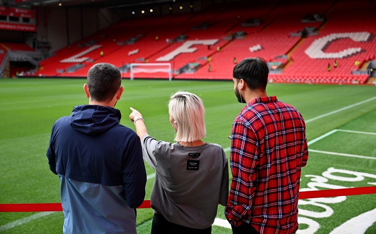 Three people observing the football pitch at Anfield, with red stadium seats in the background.