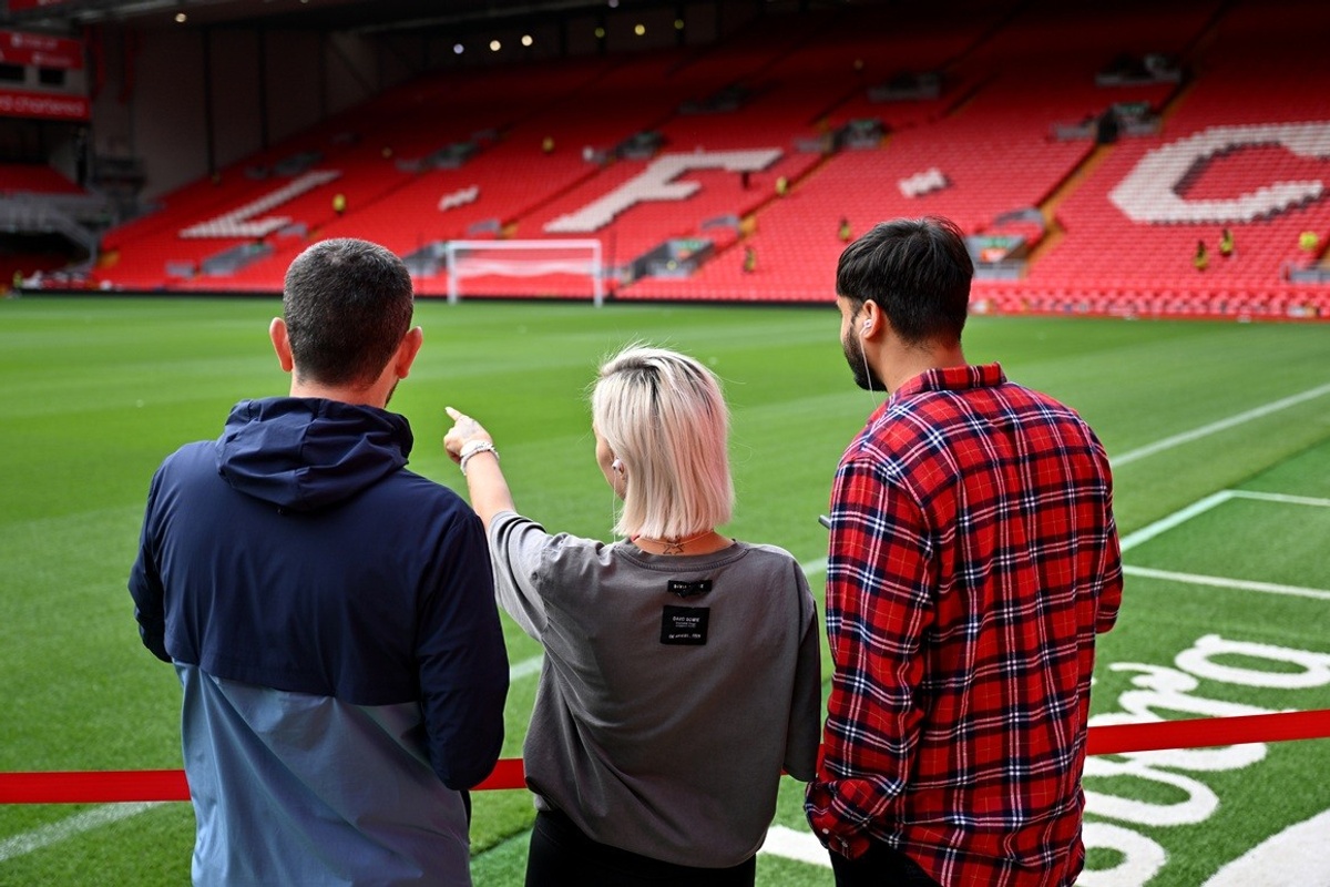 Three people observing the football pitch at Anfield, with red stadium seats in the background.