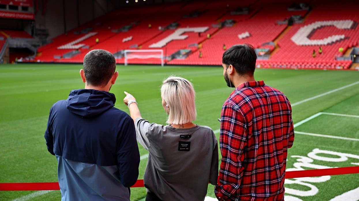 Three people observing the football pitch at Anfield, with red stadium seats in the background.