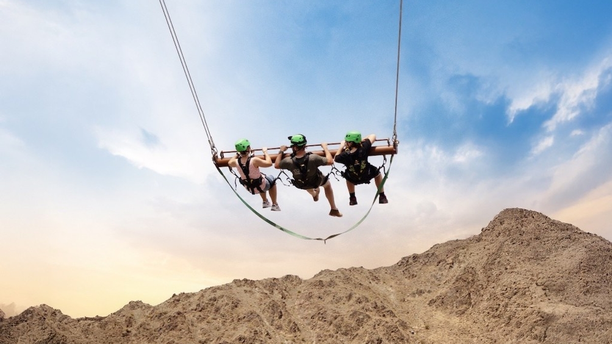 Three people on a giant swing above rocky desert landscape under a blue sky.