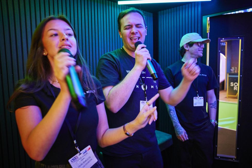 Three people singing with microphones in a neon-lit room.