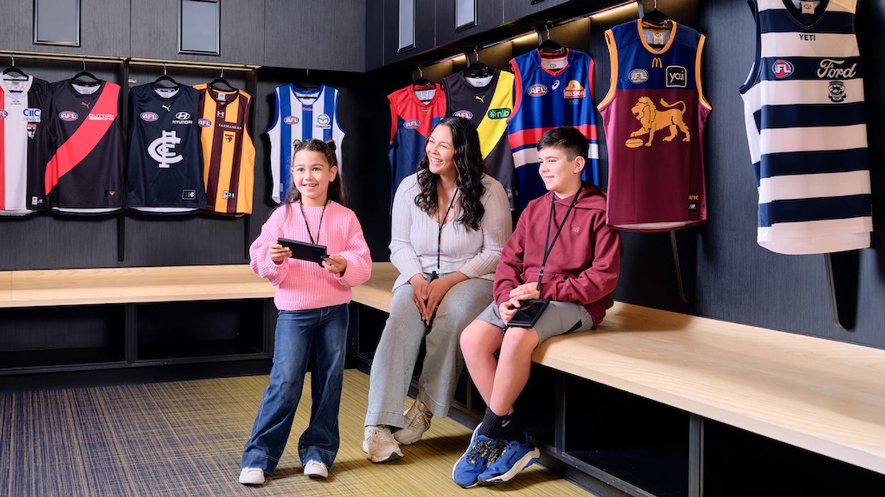 Three people sitting in a sports locker room with AFL jerseys hanging behind them.
