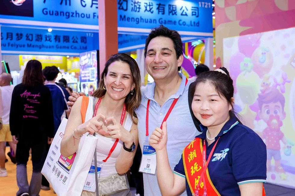Three people smiling at a gaming event, with colorful booths in the background.