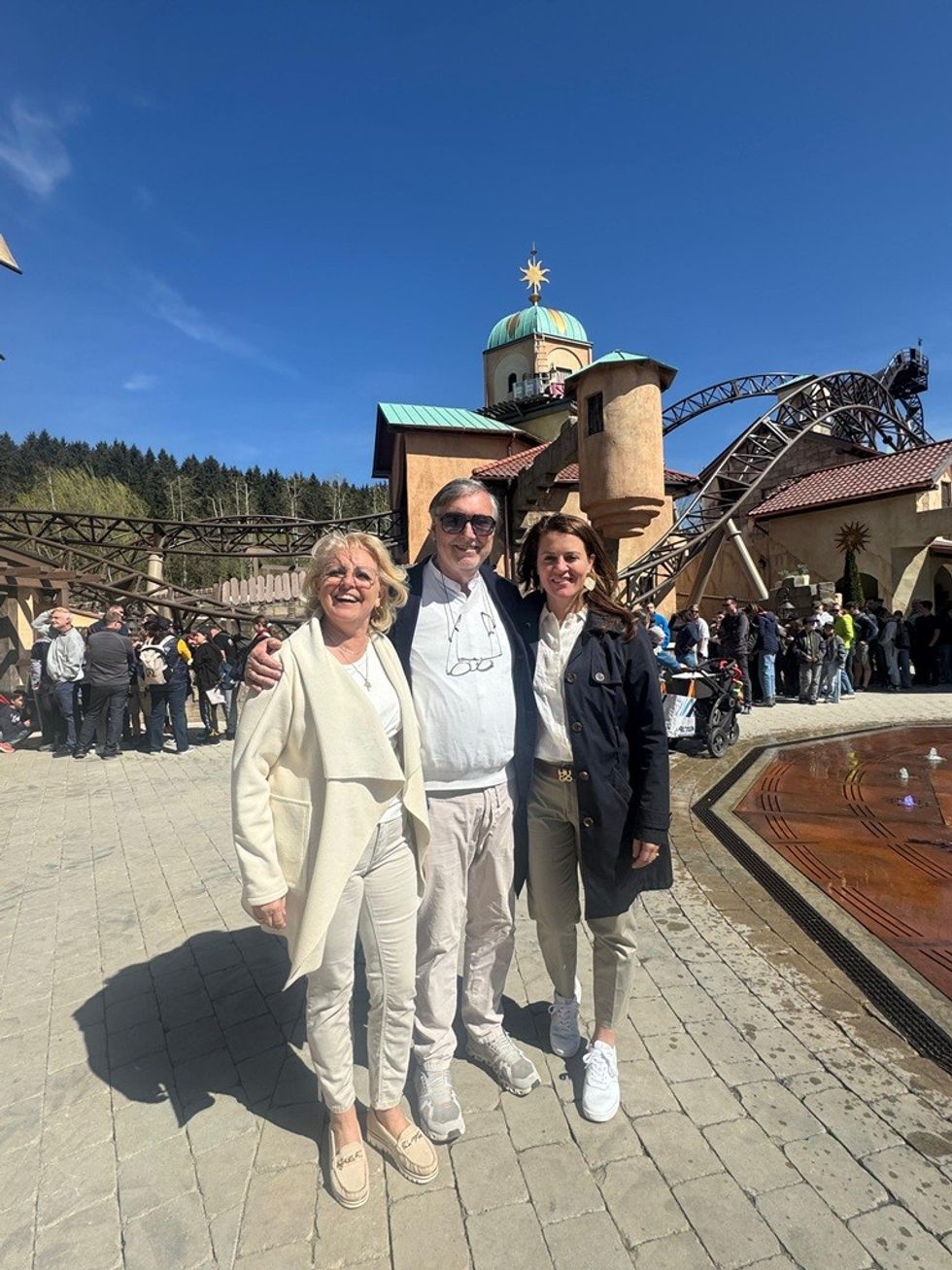 Three people smiling at a theme park with a roller coaster in the background.